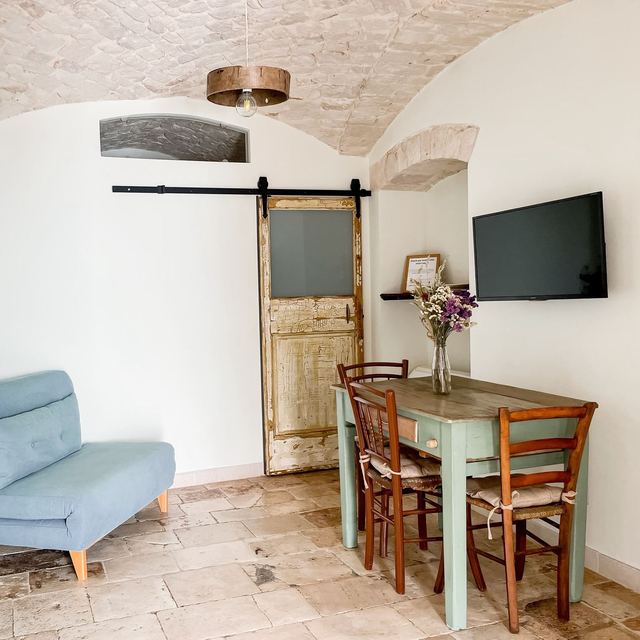 Zona living del Girasole con divano azzurro, tavolo verde salvia, TV e porta scorrevole in legno d'epoca sotto la volta